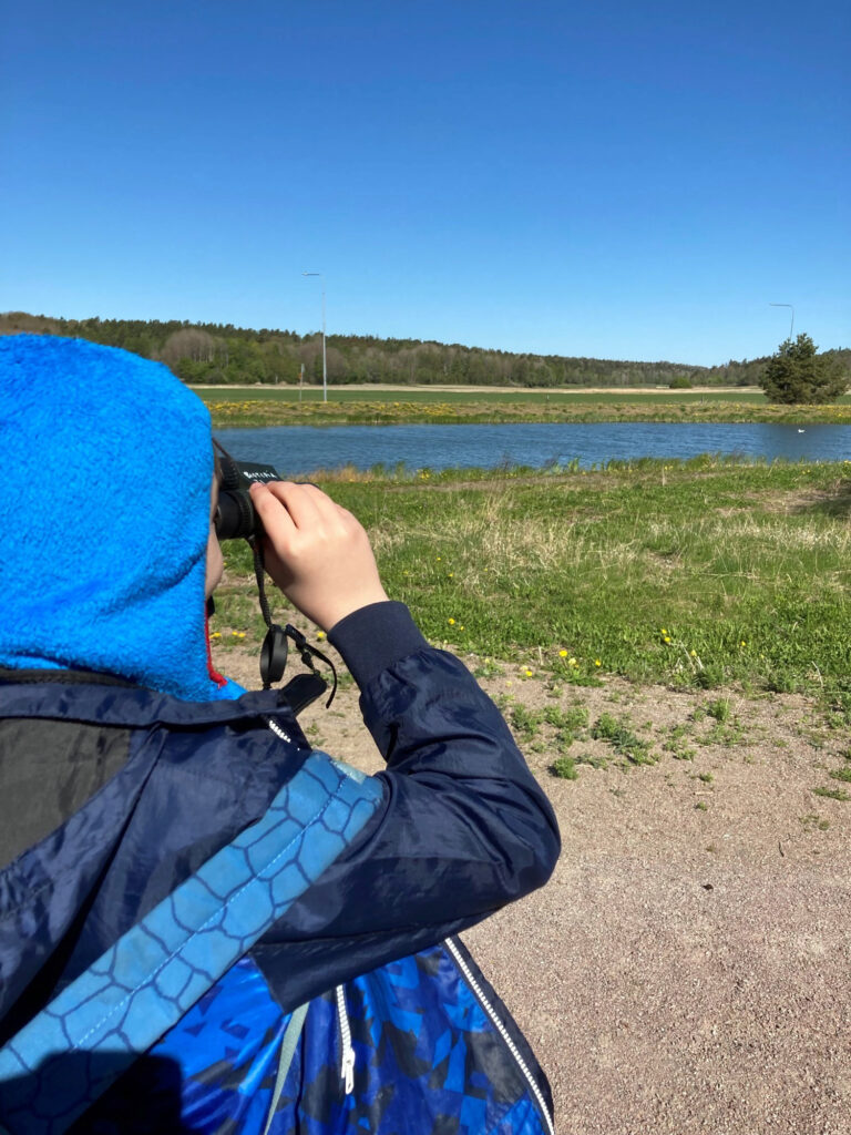 En person med blå jacka och luva tittar i en kikare mot ett vattendrag.