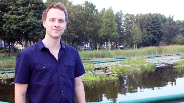 En man i blå kortärmad skjorta står framför en damm. I bakgrunden syns en skogsdunge.