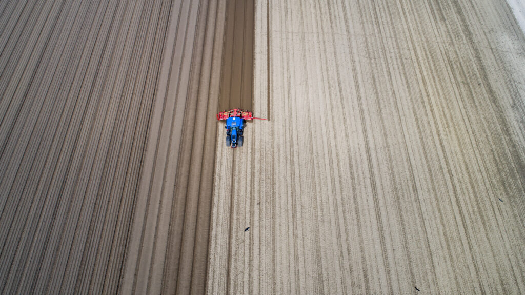 Ett flygfoto över en brun åker där en blå traktor kör med en röd plog.