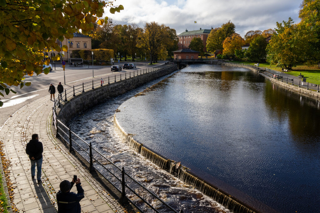 En å i en stadsmiljö med en grönskande park på ena sidan och en trottoar och ett torg på andra sidan. I ån finns en nedsänkt ränna där vatten faller ned från huvudfåran. I rännan forsar vattnet fram, i kontrast mot den stilla vattenspegeln i ån. Några personer går på trottoaren. En man håller upp sin mobiltelefon och ser ut att ta en bild på ån. I bakgrunden syns en stor röd byggnad med en svensk flagga på taket. Det är en solig dag där träden i parken står i gula och orangea höstfärger.