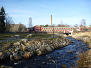 Ett vattendrag till höger med promenadstråk och en picknickplats till vänster. I bakgrunden syns en röd fabriksbyggnad.