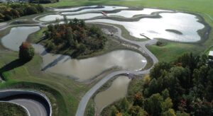 Bilden visar en parkmiljö med ett grönskande fält, skogspartier och dammar fyllda med vatten.