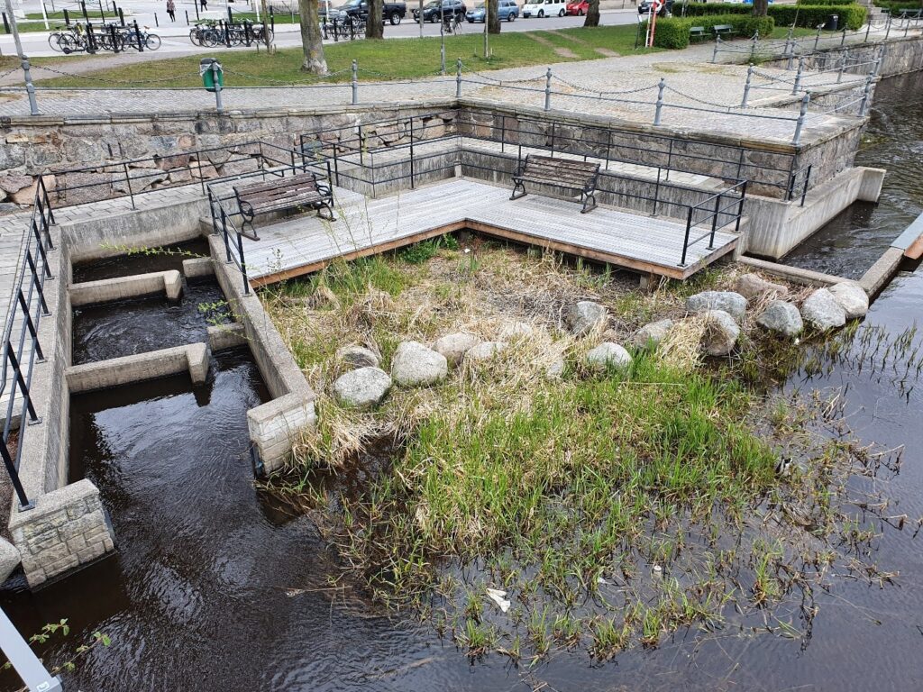 Fiskvägen vid Kanslibron i Örebro, med räcken, en gångbro och parkbänkar vid vattnet.