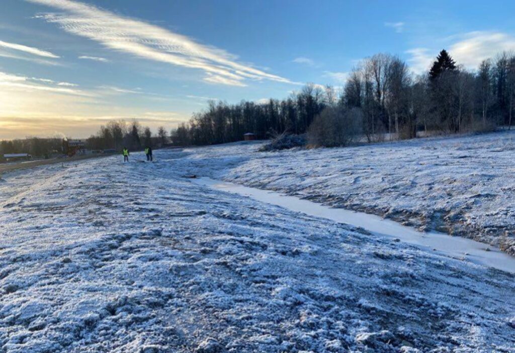 Bilden visar ett fält i vinterlandskap. Till höger skymtar en bäck med is och i bakgrunden syns tre människor.