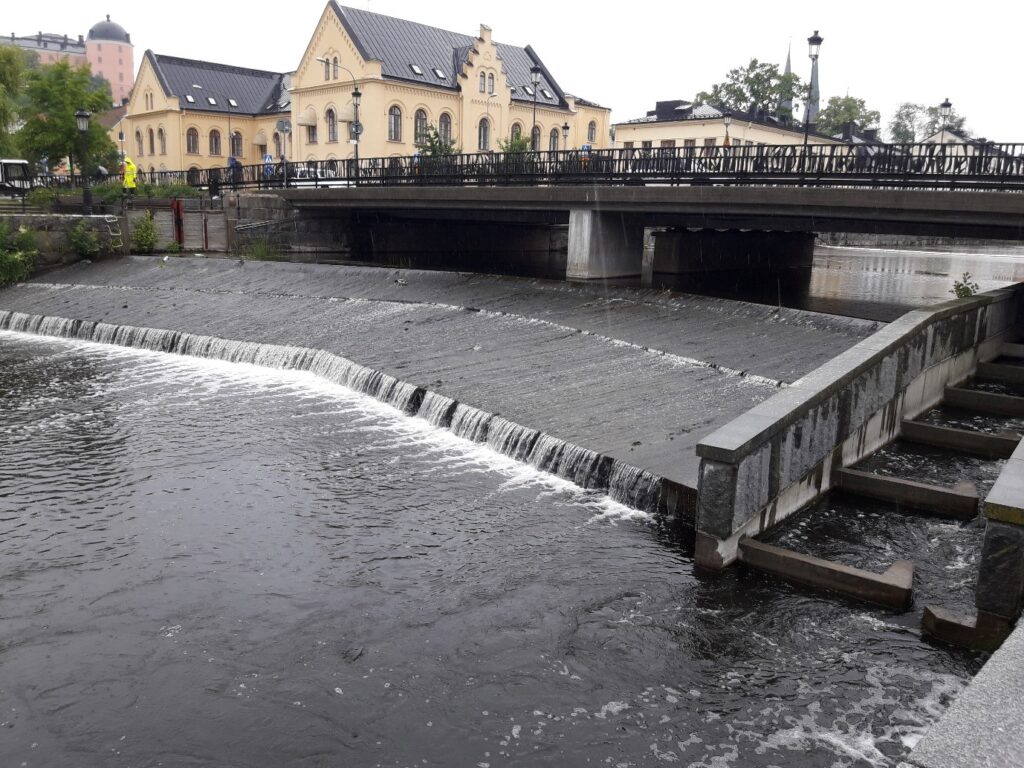 Bro med byggnader i bakgrunden och ett litet vattenfall i förgrunden. Till höger i bilden syns fisktrappan.