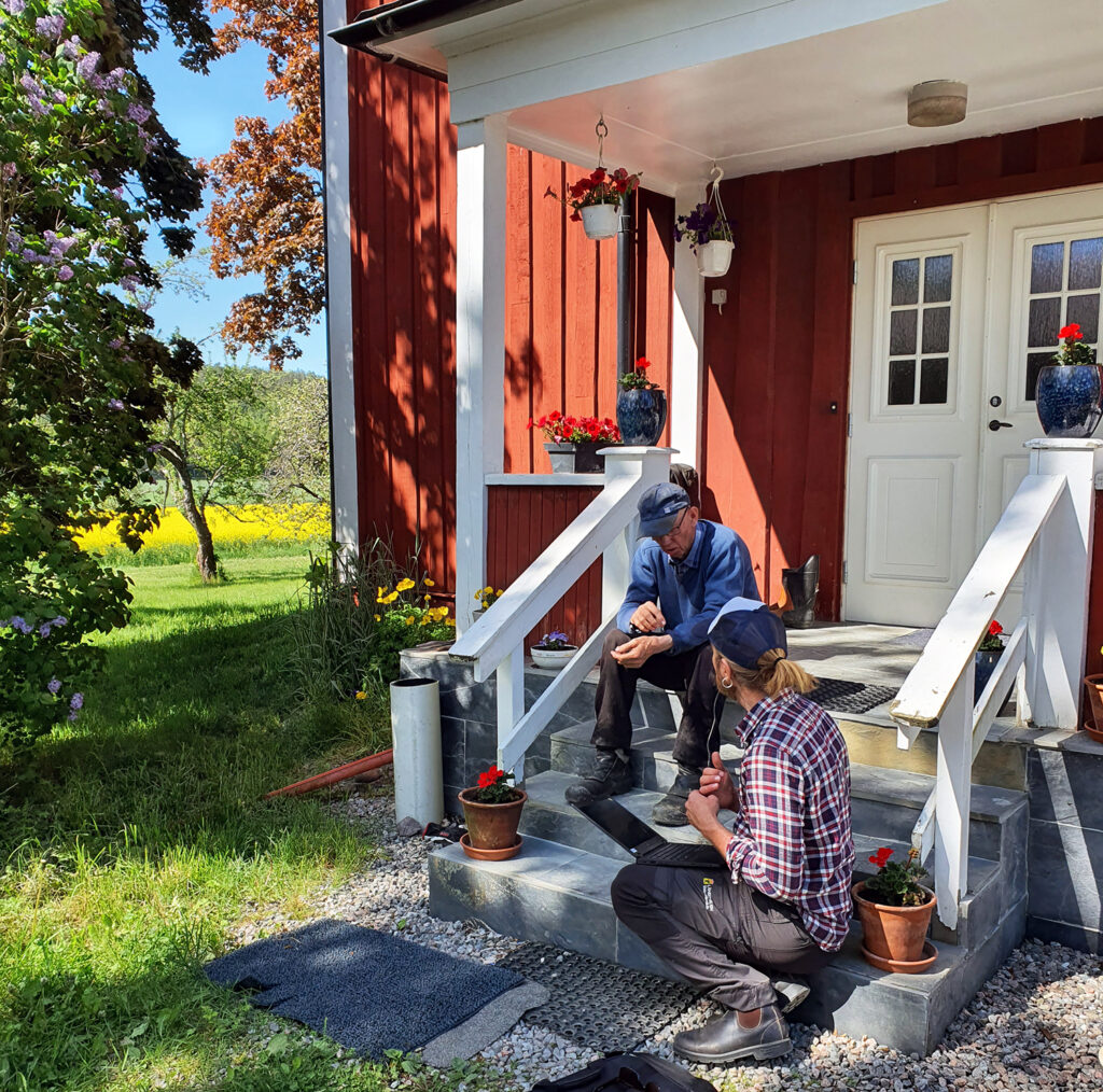 Två män sitter på trappen till ett rött hus i lantlig miljö.