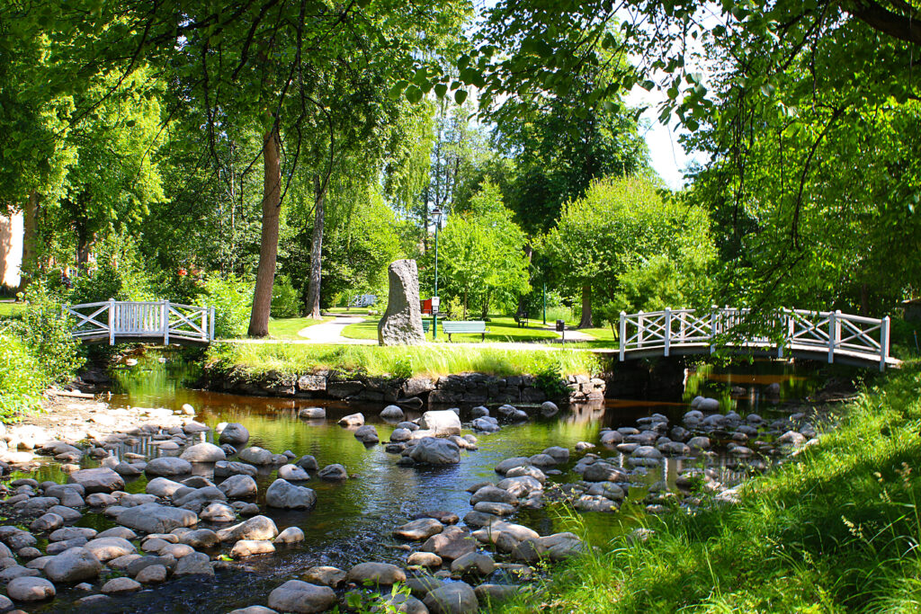 Å med stora stenar och grönskande strandkant. I bakgrunden syns gångbro och en parkmiljö.