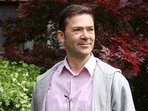 En man med kort brunt hår, rosa skjorta och grå luvtröja.