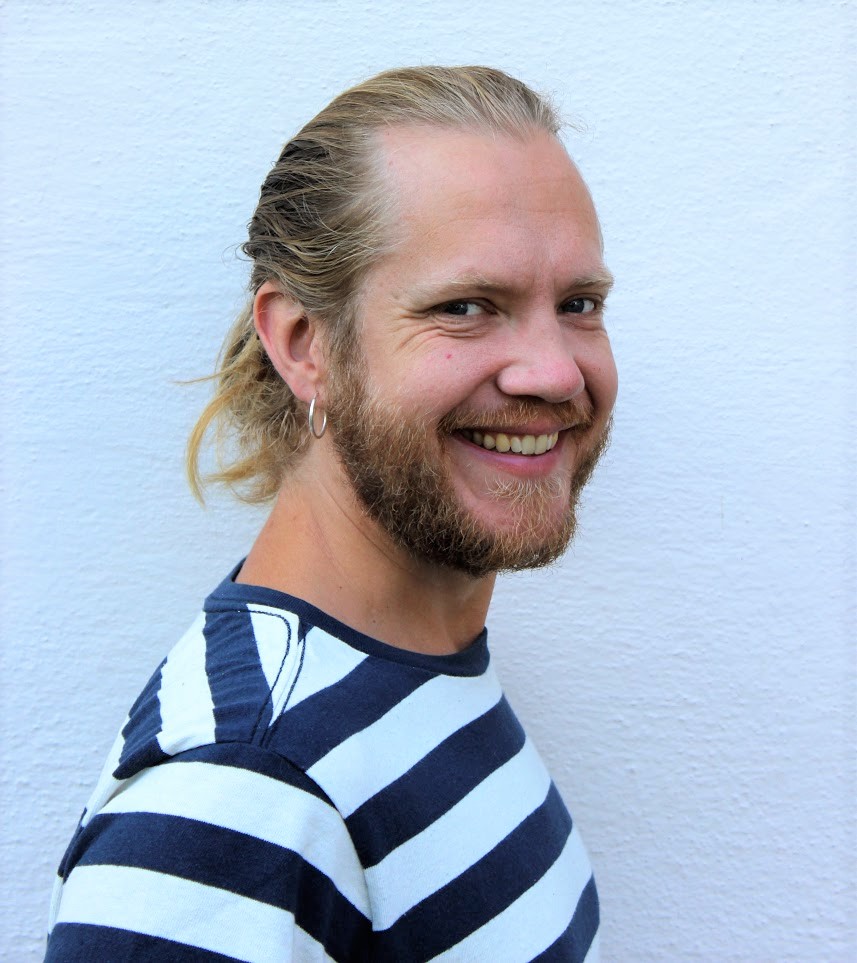 En man med långt ljust hår uppsatt i hästsvans och blå- och vitrandig t-shirt.