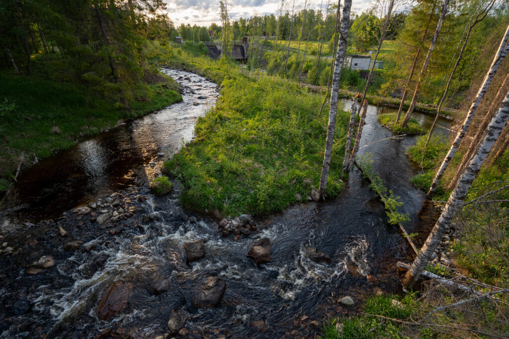 En bäck slingar sig fram genom landskapet och forsar över stora stenar i förgrunden. Det är en solig dag med grönskande buskar, träd och en äng i bakgrunden. Vid vattnet står björkar och granar. Längre ned syns en stor byggnad, en kraftstation.