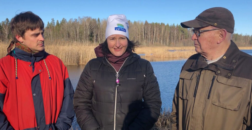 Tre personer som står vid en sjö med vass.