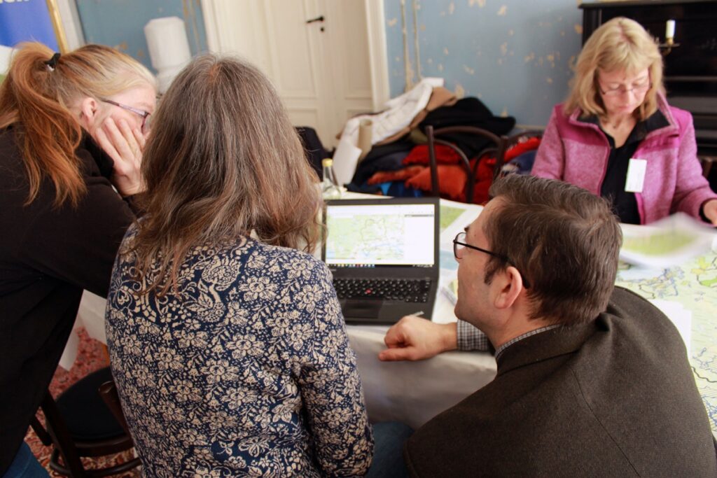 Två kvinnor och en man sitter vid ett bord och tittar på en skärm. I bakgrunden skymtar en kvinna i rosa jacka, som studerar en karta.