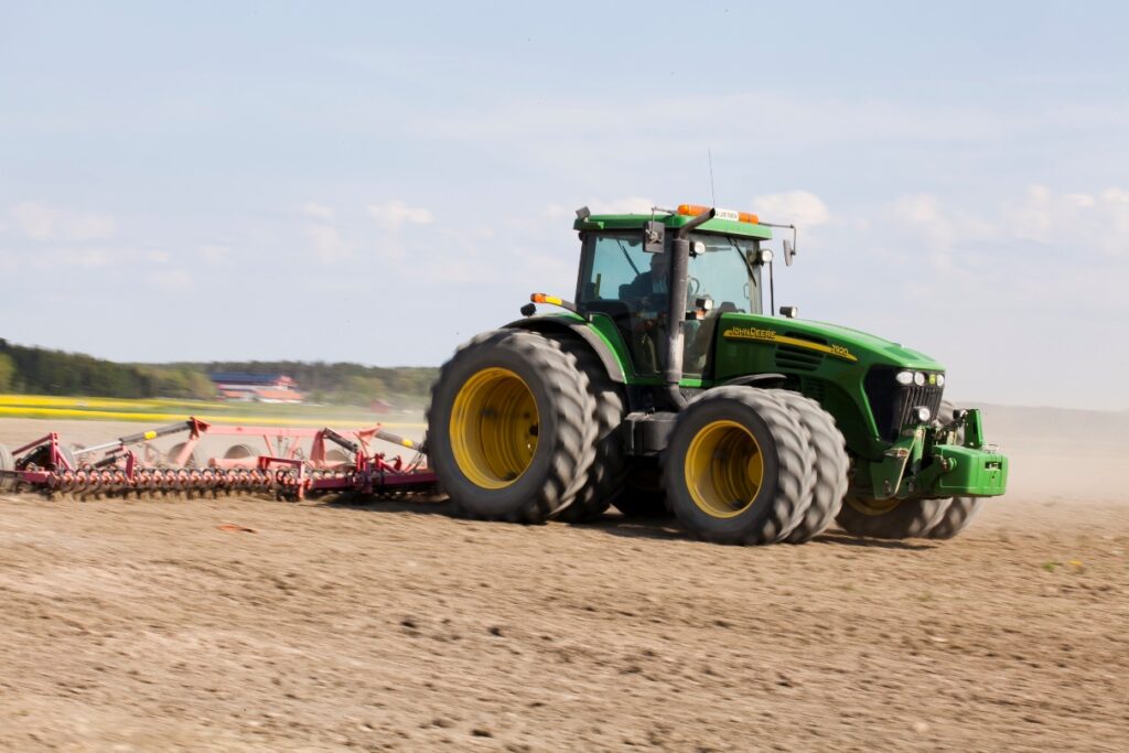 En grön traktor som drar en röd harv över en åker en sommardag