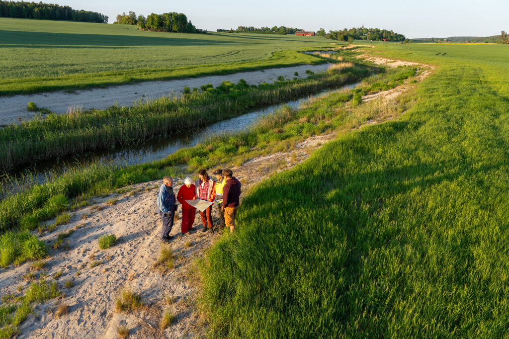 Tre män och två kvinnor står tillsammans vid en å som rinner genom ett jordbrukslandskap. En kvinna i mörkt hår och röda kläder håller i en stor karta som de andra tittar på. Landskapet består av gröna fält, med skog i bakgrunden. Man ser även en ladugård längre bort där fälten tar slut.