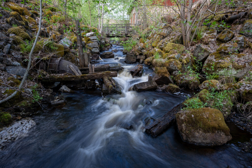 Ett vattendrag som forsar fram över stora stenar i ett grönskande skogslandskap. I bakgrunden syns en bro.