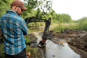 En man med keps vid ett vattendrag där en grävskopa gräver.