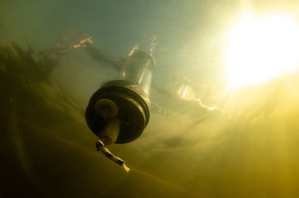 Ett rör med metallbotten i närbild under vattnet. Ovanför vattenytan skymtas en båt. Vattnet är grönbrunt och solens strålar tränger ned i övre delen av bilden.