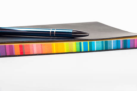 En svart skrivblock med färgade sidor och en mörkblå penna.