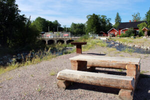 Picknickmöbel med grillplats bredvid ett vattendrag med stenar. I bakgrunden syns en vägbro och röda hus.